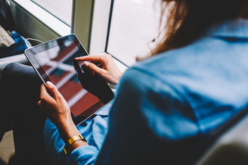 Cropped back view of young woman choosing music application on website touching with finger on display of modern touch pad connected to public transport wifi.Female passenger with tablet in hands