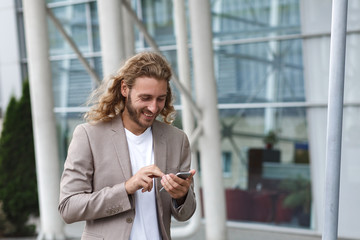 Portrait of handsome curly businessman in casual wear holding smartphone and smiling.Successful manager using mobile phone apps,texting message,browsing internet,looking at phone near business center.