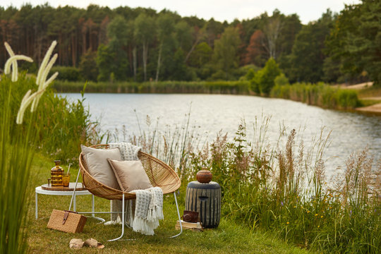 Stylish Composition Of Outdoor Garden On The Lake With Design Rattan Armchair, Coffee Table,  Plaid, Pillows, Drinks And Elegant Accessories. Summer Vibes.