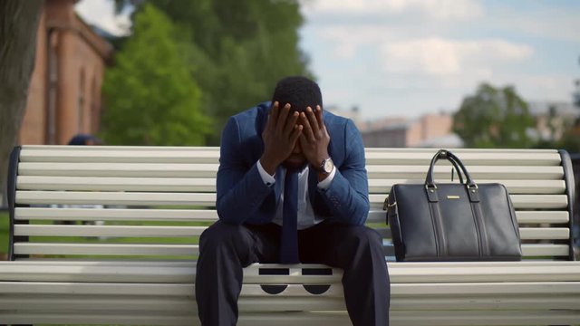 African businessman feeling stressed and depressed sitting on bench outdoors.