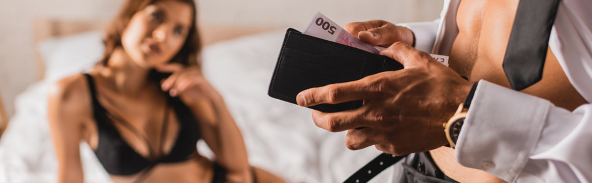 Panoramic shot of muscular man holding wallet and money near woman in bra on bed