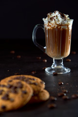 Close up of a glass cup of coffee with whipped cream and chocolate on it, chocolate chip cookies...