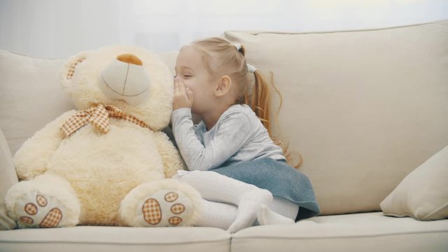 4k slowmotion video of little girl sitting on the sofa, talking with her teddy and showing silent sign.