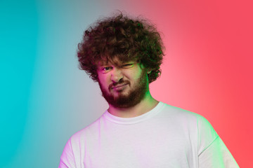 Disguasted. Caucasian young man's portrait isolated on gradient studio background in neon light. Beautiful male model in casual style. Concept of human emotions, facial expression, youth, sales, ad.