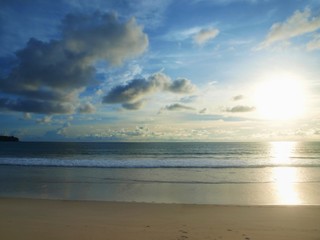 Sky with white-gray cumulus clouds and sea at sunset. Reflection of the sun moving to the horizon on the water surface. Open ocean, weak waves roll on the yellow sand of an empty beach