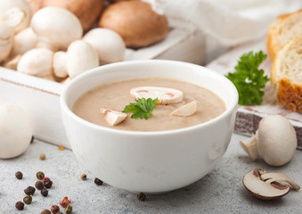 White bowl plate of creamy chestnut champignon mushroom soup on white kitchen background and box of raw mushrooms and fresh bread.