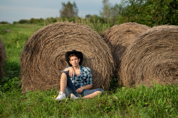 Naklejka na ściany i meble A young man in a plaid shirt, a cowboy hat with a straw in his mouth sits near a roll of hay and looks into the distance. Farmer man sitting by the hay.