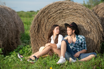 Naklejka na ściany i meble Brother and sister sitting together by the haystack. A young man with a straw in his mouth sits near a roll of hay and looks into the distance. Farmer man and girl sitting by the hay.