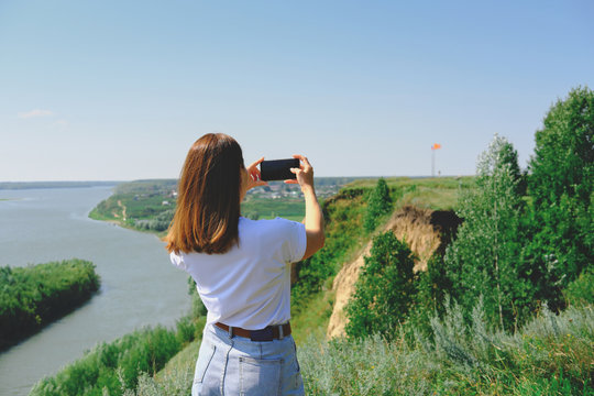 beautiful woman taking a picture of a landscape on a smartphone. back view, unrecognizable person.woman wearing white t-shirt and jeans photographing red flag with a phone camera. travelling concept.