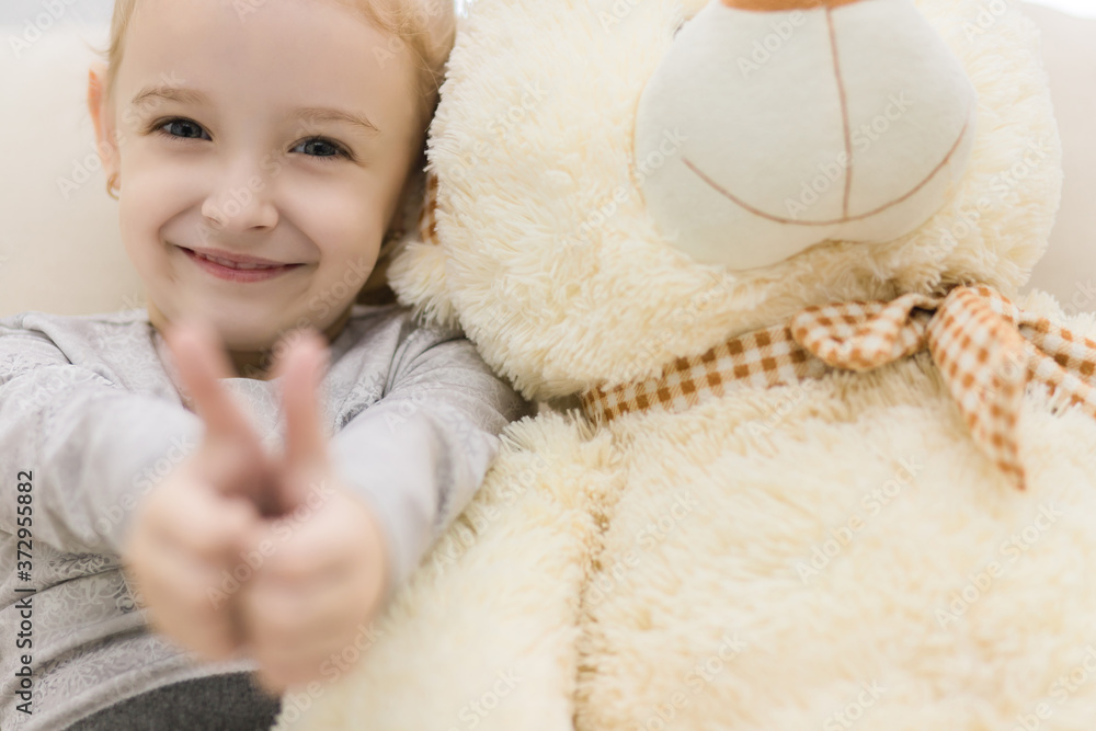 Wall mural photo where little girl showing two thumbs up near white teddy bear.