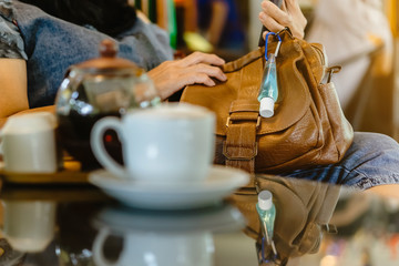 Mini portable alcohol gel bottle to kill Corona Virus(Covid-19) hang on a brown leather shoulder bag with hot coffee in cafe.New normal lifestyle.Health care concept.Selective focus on alcohol gel