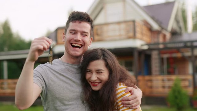 Young married couple or family buy a new house. Happy man and woman or owners shake key in air and hug each other in front of their newly bought home. Excited clients purchasing estate or apartment