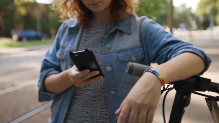 LGBT woman stands by rented electric scooter using smartphone and wearing rainbow bracelet - Powered by Adobe