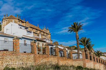 Brick wall with pillars and wrought iron bars in a street on a hill with palm trees with houses and the Royal Collegiate Church of Santa María la Mayor in the background, sunny day in Antequera, Spain