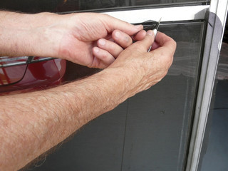 Trimming Wire Screen Door with Knife. A homeowner is using a knife to trim wire mesh after inserting spline in a project to replace a damaged panel in a sliding garage screen door with a metal frame.