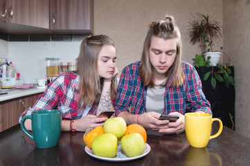 Young couple man and woman with long hair are sitting at the kitchen table in the kitchen and using their smartphones while buying food on the internet