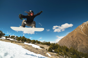 Stylish young girl snowboarder does the trick in jumping from a snow kicker against the blue sky clouds and mountains in the spring.