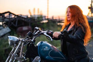 Obraz na płótnie Canvas Curly red-haired woman in a black leather jacket sits on a motorcycle at sunset. Portrait of a serious girl driving a bike.