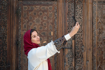 Young muslim woman taking selfie with mobile phone in traditional clothing with red headscarf
