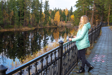 Fototapeta na wymiar A blonde woman in a jacket stands on the embankment of the river in the autumn forest among tall pines.