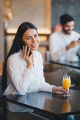 A young woman is sitting at a table in a restaurant, talking on a mobile phone, smiling and drinking orange juice, young gentleman is sitting behind her and looking at the phone