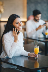 A woman is sitting at a table in a restaurant, talking on a mobile phone, smiling and drinking orange juice, young gentleman is sitting behind her and looking at the phone