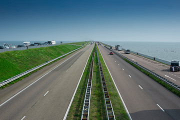 Straight asphalt road. Major dam with causeway,  green grass and cars travelling the road. Sea, blue sky as background.