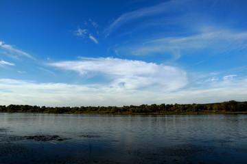 Wide view of a calm lake under a blue sky with cirrus clouds. Tranquil natural landscape with forested shoreline and atmospheric reflections on water