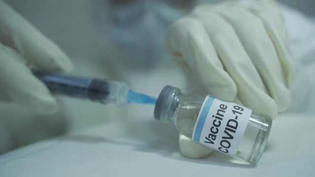 human wearing protective PPE kit carefully drawing out covid 19 vaccine from vial and filling into syringe in horizontal position. close up shot.