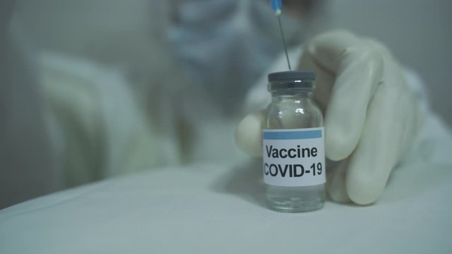 human wearing protective PPE kit inserting syringe in covid 19 vaccine vial and preparing to withdraw.