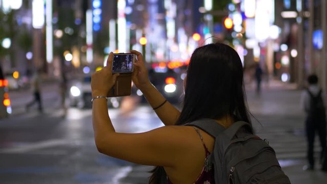 A young female tourist on the streets of Tokyo at night in summer taking photos on her smartphone.
