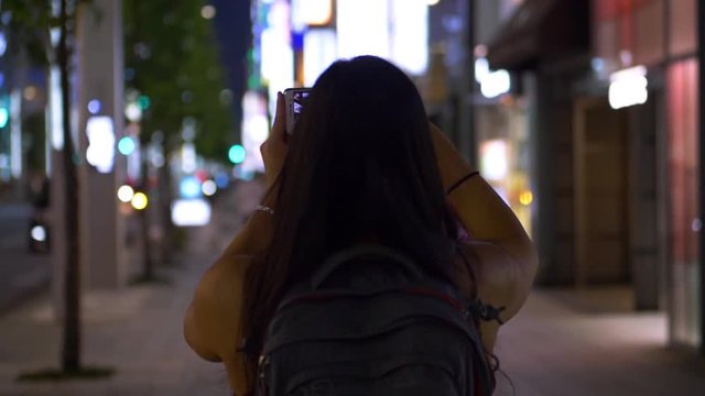 Slow Motion shot from the back of a young female tourist walking and taking photos of the streets of Tokyo at dusk.