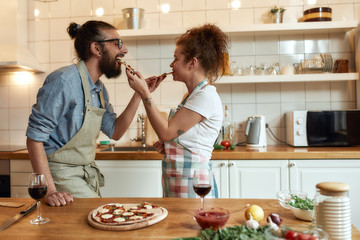 Eat and enjoy. Young man and woman in apron feeding freshly baked pizza to each other while standing in the kitchen. Love, relationships concept