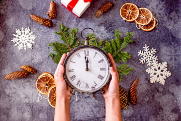 Fototapeta na wymiar Christmas clock showing midnight in women's hands on a dark background, decorated with fir branches, cones, dried oranges. Happy new year background with vintage clocks and hands. Copy space, top view