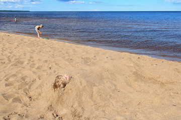 blond boy is buried in the sand on the beach.