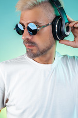 Confident bearded hipster young man wearing sunglasses listening musicin stylish headphones on cyan background.