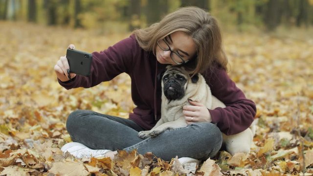 Teenager girl making selfie with funny cute pug dog. Pug dog posing for the camera. Evening autumn park, forest. Yellow autumn leaves. Teen photography hobby concept. Taking photos on a smartphone