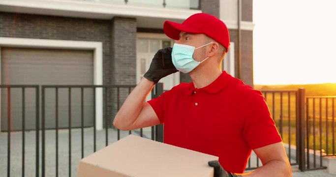 Caucasian delivery man in medical mask, gloves and hat standing outdoor with carton parcels and talking on cellphone. Male courier in uniform speaking on mobile phone and searching adress. Delivering.