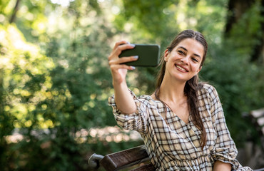 A young woman is sitting on a park bench and taking a picture of herself on her mobile phone