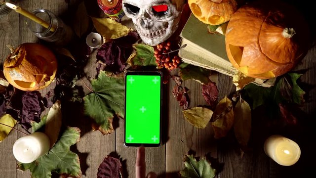 Using a phone with a green screen on background of Halloween decorations. Top view decorations for all saints day and smartphone. Mystical tradition in harvest autumn desk decor, copy space template.