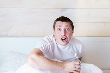 Drunk rowdy man with angry face, bottle of alcohol sits drinking in bed in wooden bedroom. Man with...