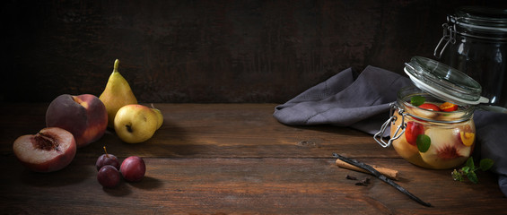 Peach, pear and plums, fresh fruits and preserved with cinnamon and cloves in a glass jar on dark rustic wood, moody vintage style in panoramic banner format, copy space, selected focus