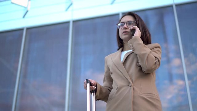 A young woman stands against the background of a business center and speaks on the phone. Girl in a coat with a suitcase closeup.