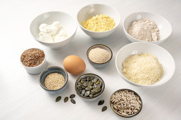 Ingredients for a protein bread with quark, oat bran, lupine flour, almond and various seeds in bowls on a white table, healthy baking for low carb or ketogenic diet