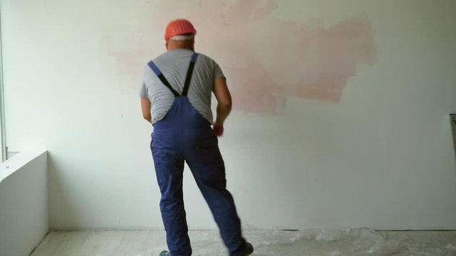 A bearded male construction worker is dressed in overalls and a protective helmet. paints the wall with a roller, general plan. Renovation concept in the room