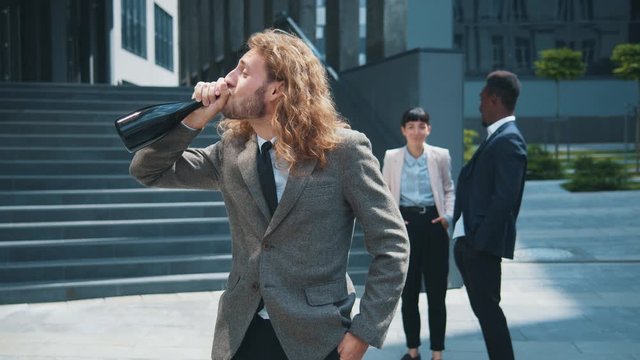 Carefree young businessman with curly hair silly smile drinking champagne celebrating job promotion. Surprised funny colleagues laugh in background.