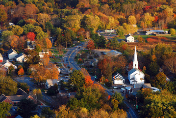 A classic New England town, with a white church with a large steeple, is surrounded by brilliant autumn foliage in an aerial view - obrazy, fototapety, plakaty