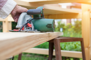 A working electric saw of green color leading with a hand along a wooden beam during construction at a summer cottage on a summer day.