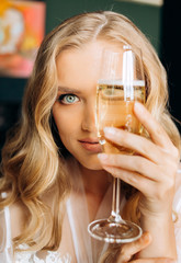 Morning of the bride. Young woman in a white coat closes part of the face with a glass of champagne.