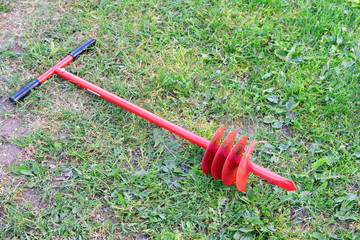 Red new metal hand drill for drilling holes lies on ground in green grass outdoors.
